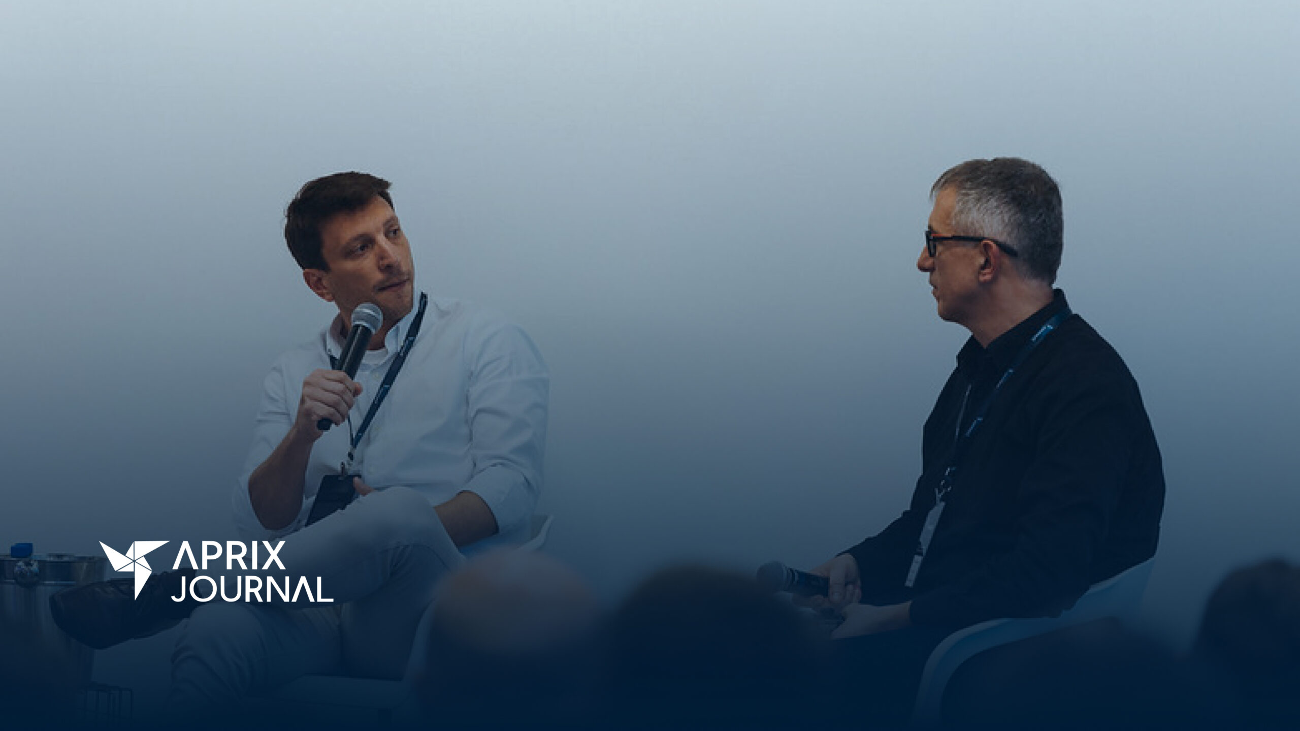 Dois homens em um palco, diante de uma plateia, estão sentados e segurando microfones enquanto conversam, durante um painel do Aprix Connect